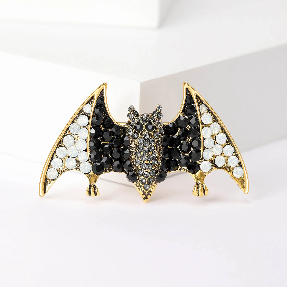Bat brooch in gold-tone metal with black and white faceted stones, upright pose on white block.
