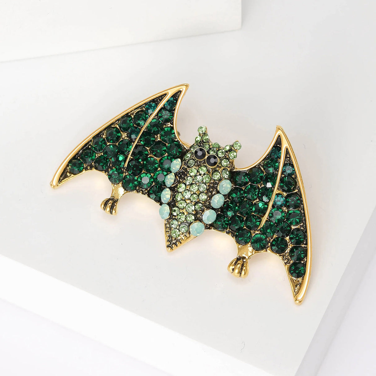 Bat brooch with dark green faceted wings, light green body stones, and gold-tone feet, angled view on white surface.