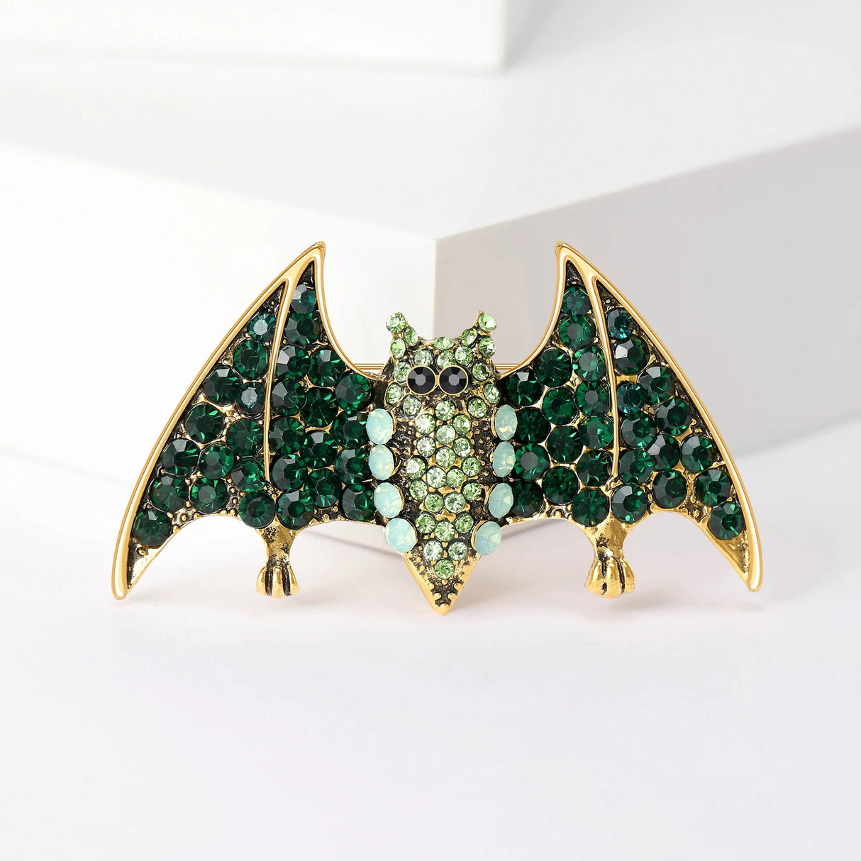 Bat brooch with gold-tone setting, deep green faceted wings, light green center stones, upright view on white block.