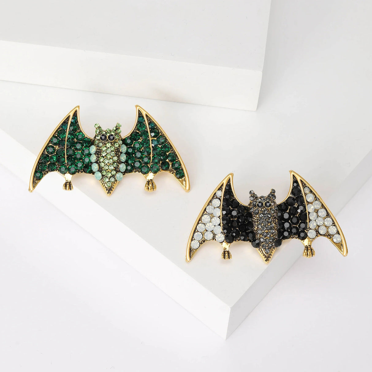 Two bat brooches with gold-tone base, one with green faceted stones and the other with black and clear stones, flat lay on white surface.