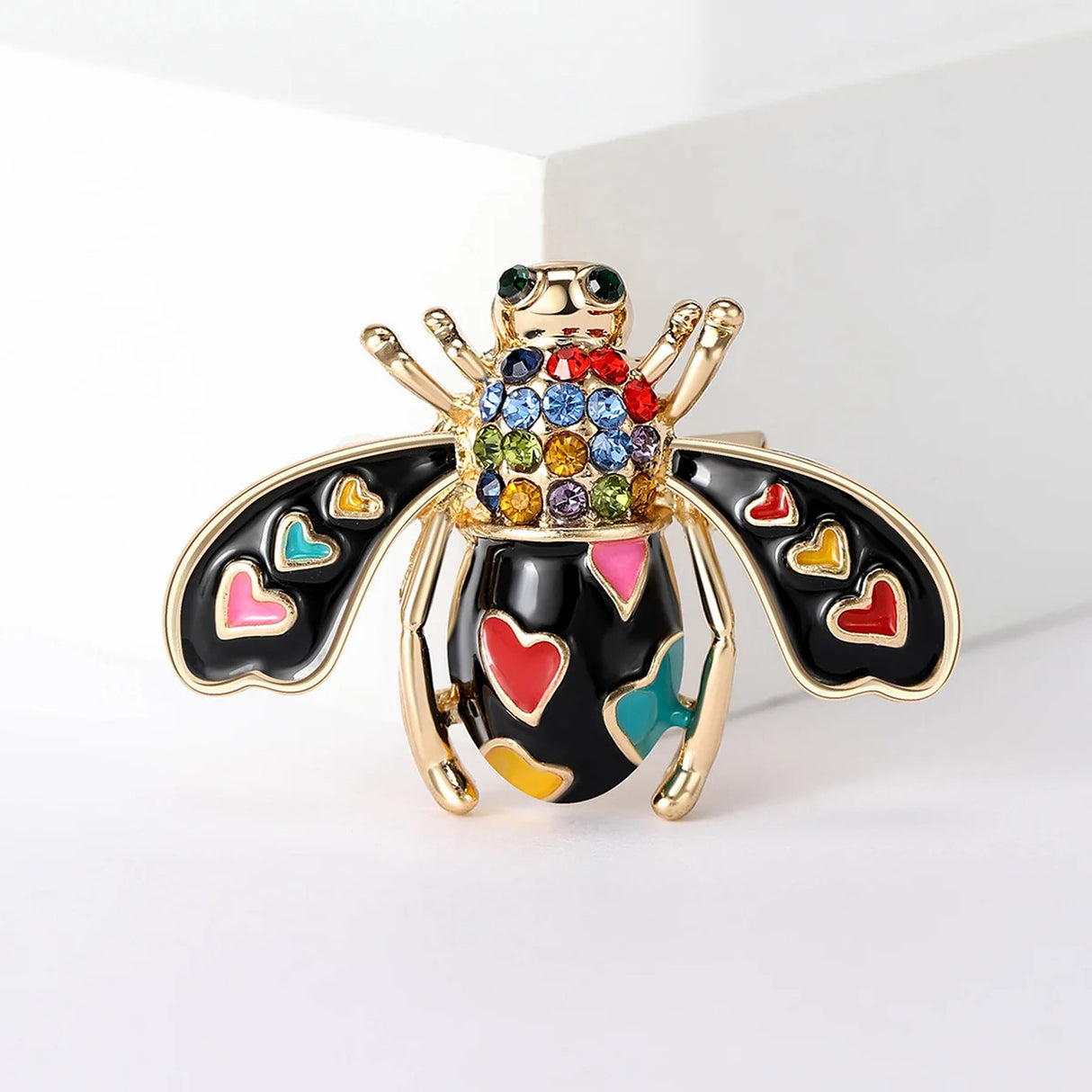 Bee brooch featuring black enamel wings with multicolor heart accents and rainbow faceted stones, front view on white.