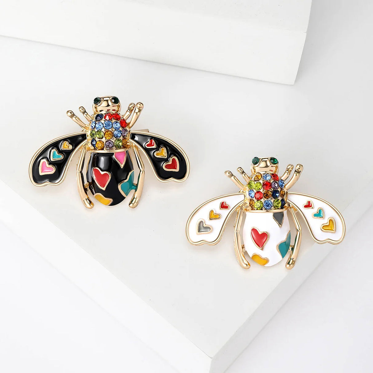 Set of two bee brooches with black and white enamel bodies, heart-shaped details, and multicolor stones, top-down view.