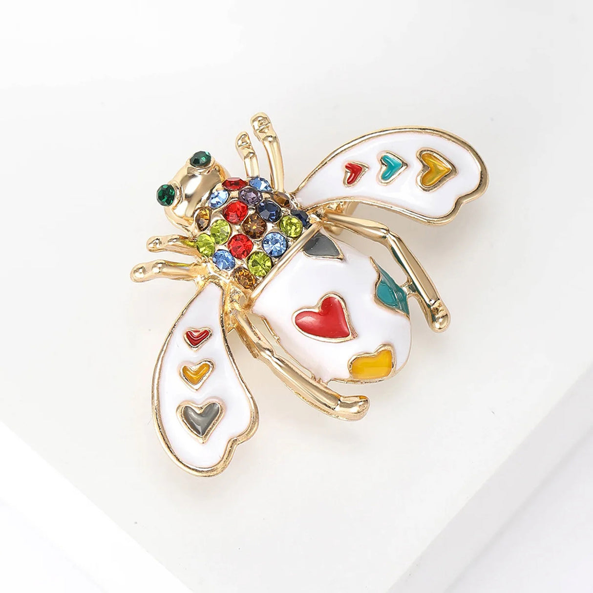 Bee brooch with white enamel wings, colorful heart accents, and multicolor faceted stones, angled view on white block.