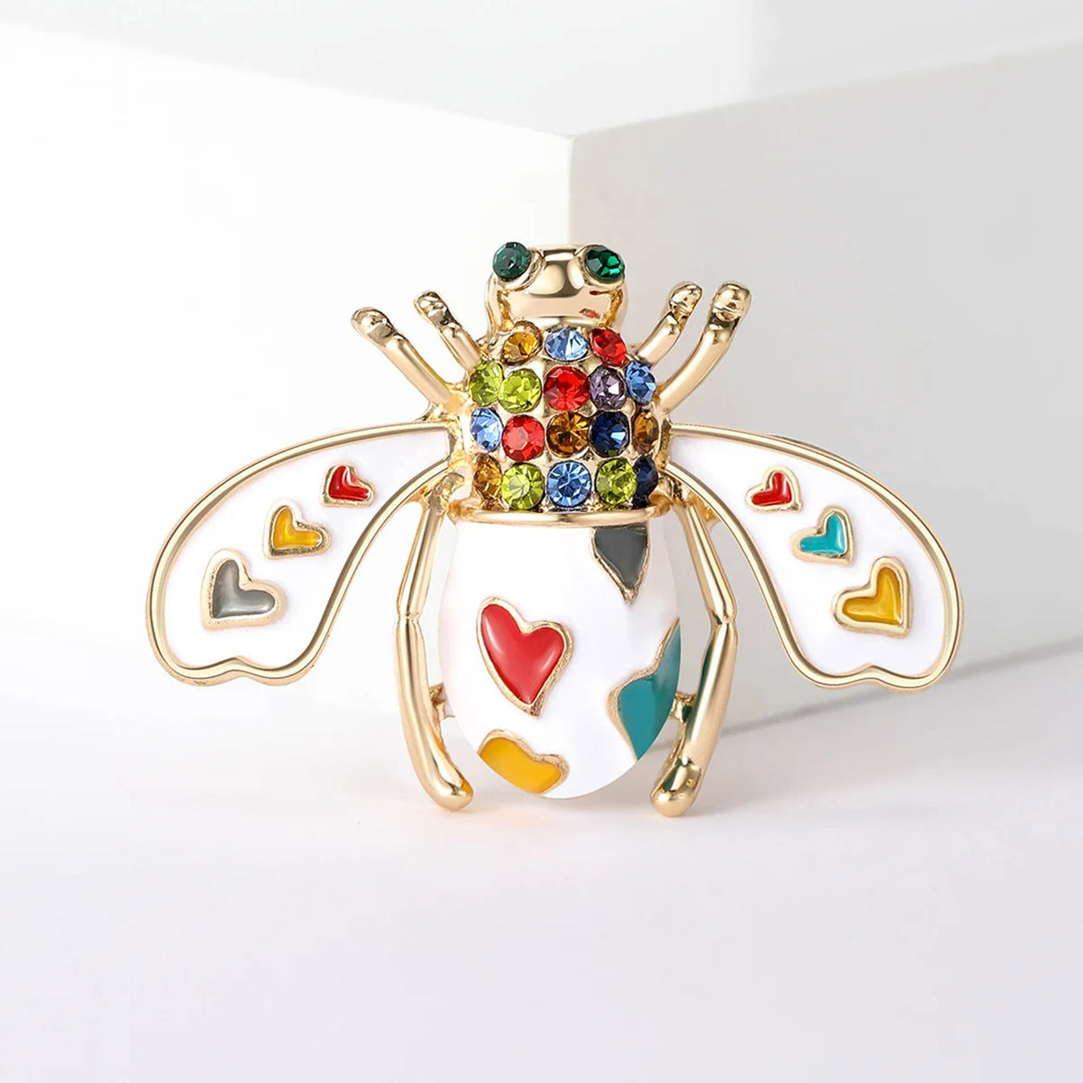 Bee brooch with white enamel wings, rainbow heart motifs, and clear stones on the thorax, front view on white.