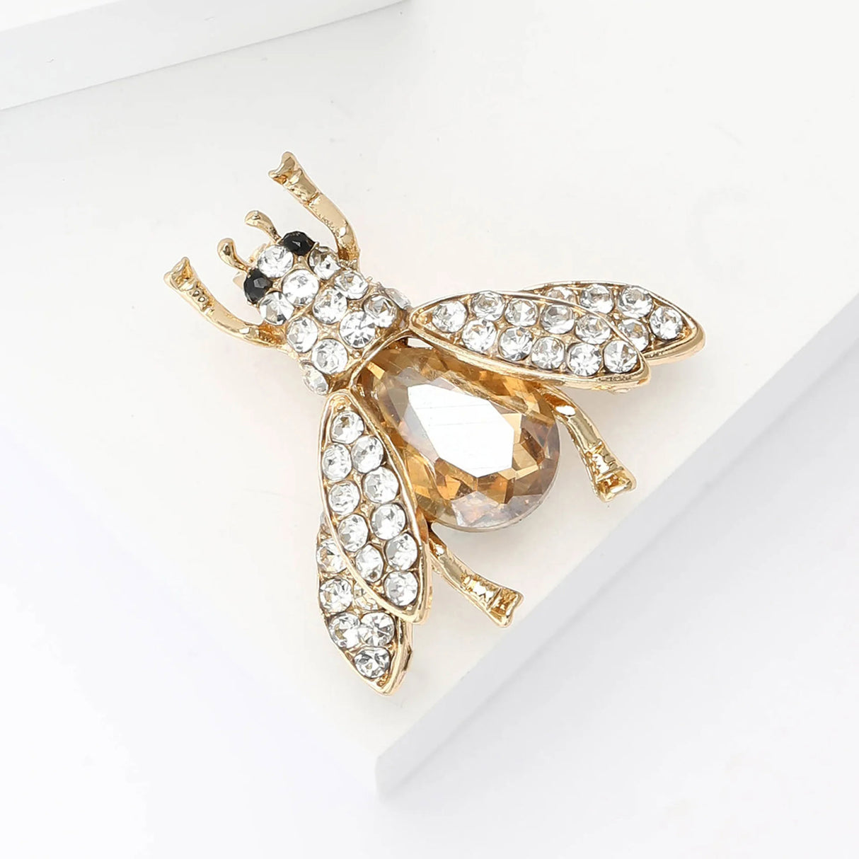 Bee brooch with gold-tone finish, clear faceted stones on wings and head, and large amber-toned body stone, angled view on white.