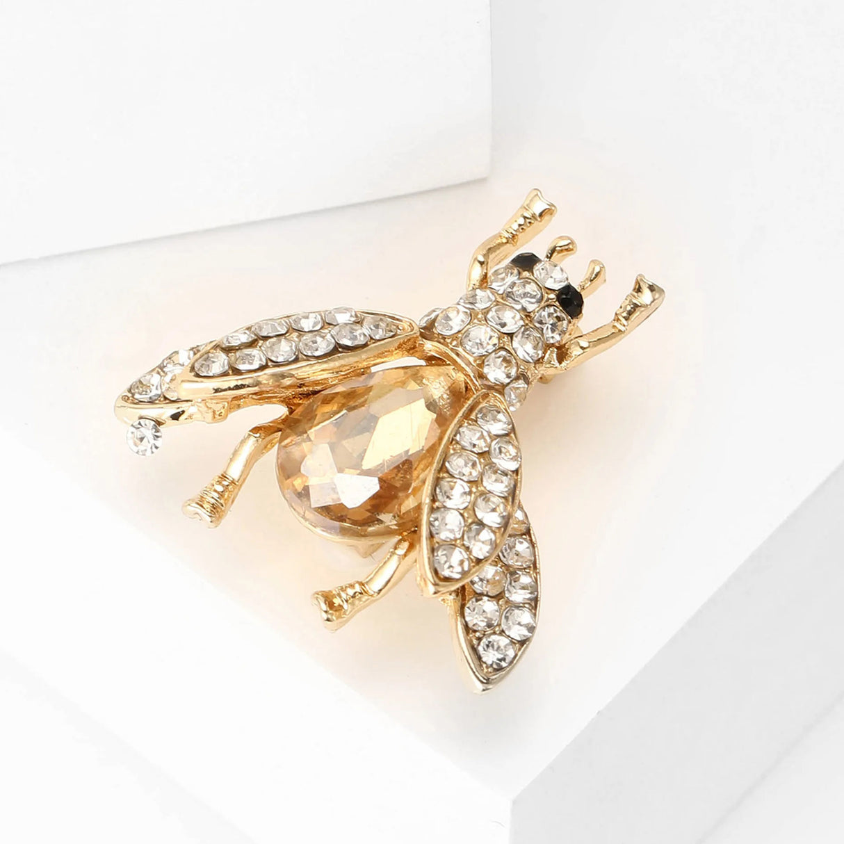 Bee brooch in gold-tone metal with clear stones on wings and head, amber-toned faceted body, angled side view on white surface.