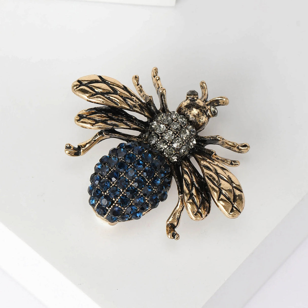 Bee brooch with textured gold-tone wings, clear stone thorax, and blue faceted stone bottom, angled view on white.