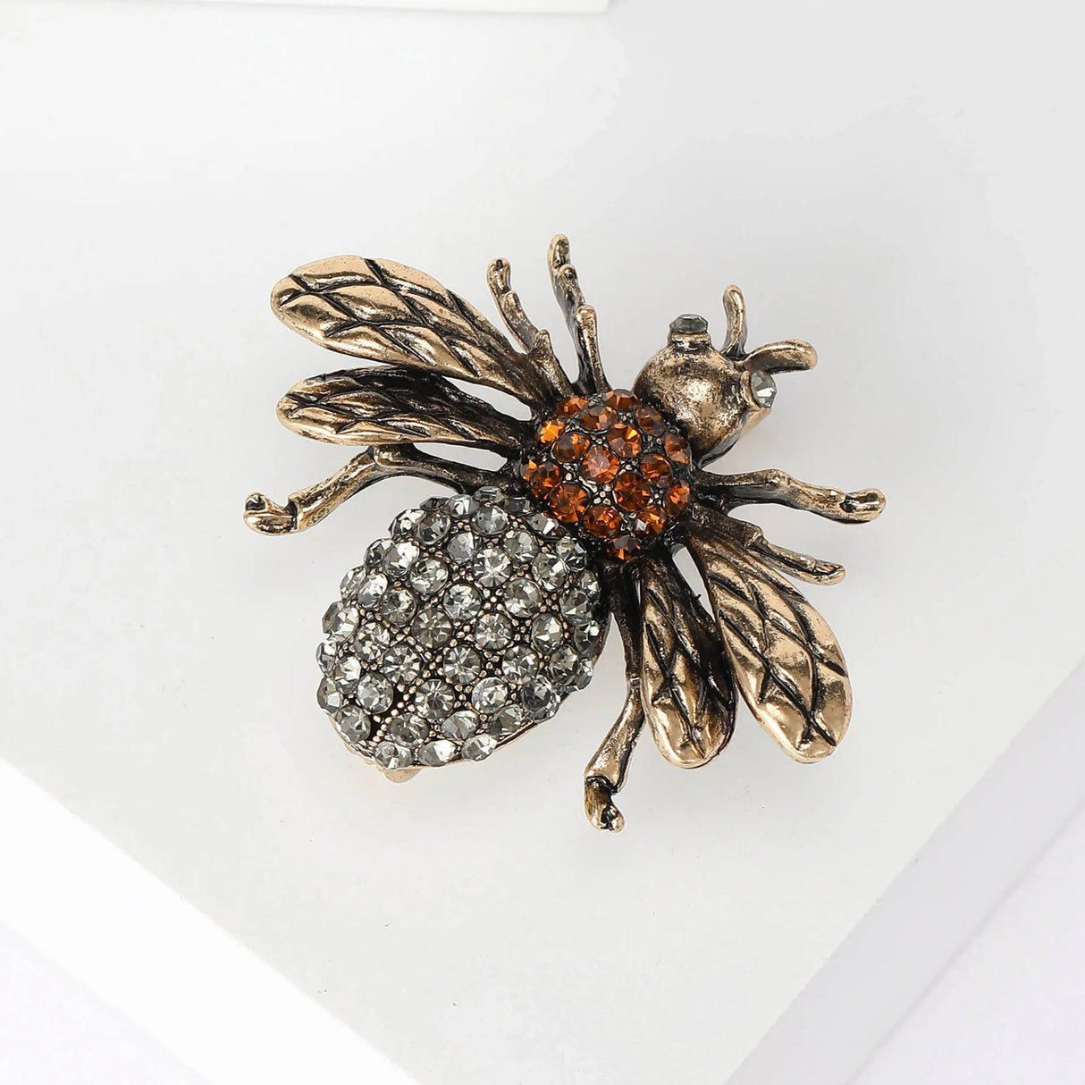 Bee brooch with gold-tone textured wings, orange-brown faceted stone thorax, and clear stone abdomen, angled view on white.