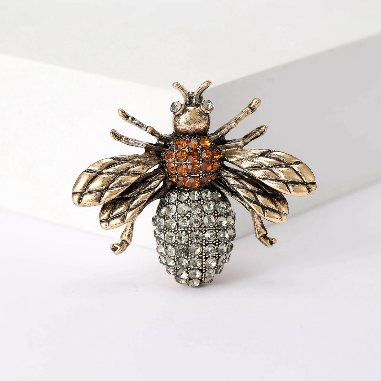 Bee brooch featuring gold-tone wings, orange-tone thorax, and clear faceted stone bottom, upright studio view.