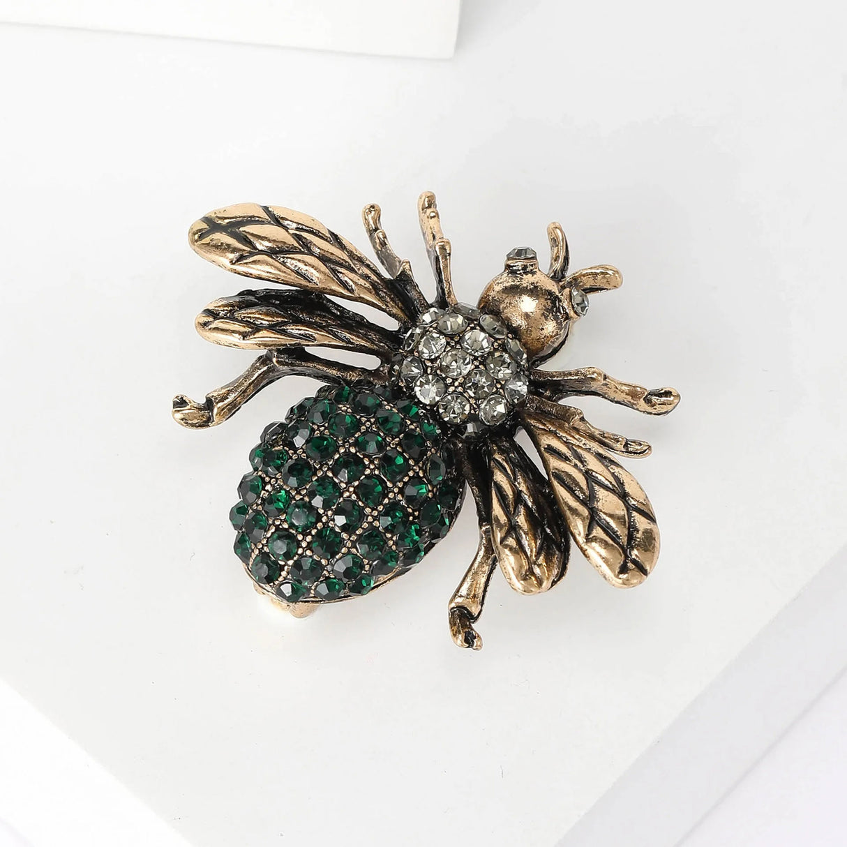 Bee brooch with gold-tone wings, clear faceted stone thorax, and green stone-covered abdomen, angled view on white.