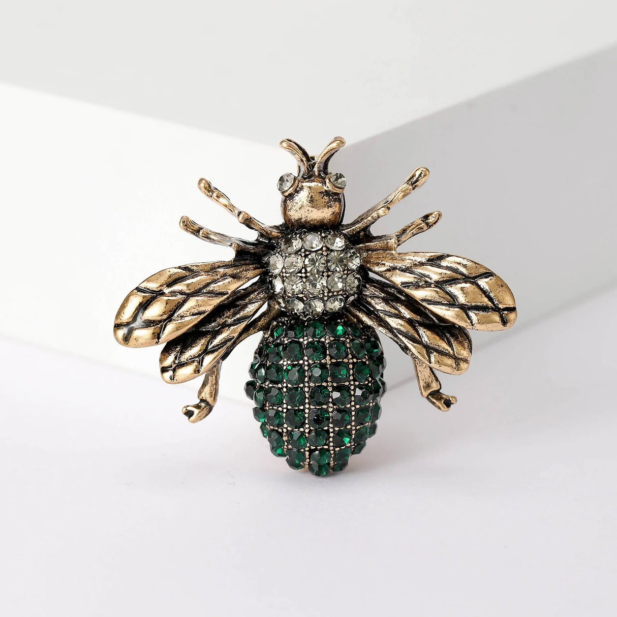 Bee brooch with textured gold-tone wings, clear thorax stones, and green faceted stone abdomen, upright view.
