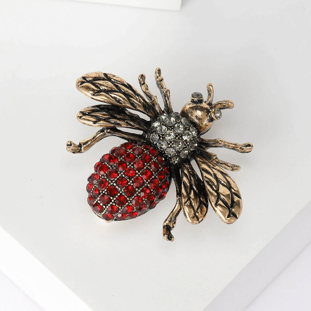 Bee brooch with gold-tone wings, clear stone thorax, and red faceted stone bottom, angled flat lay on white.