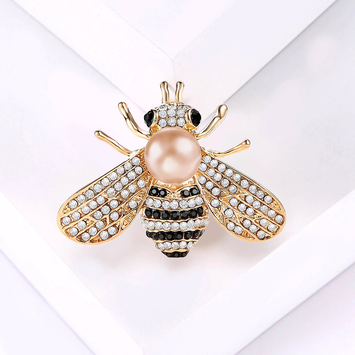 Bee brooch with gold-tone finish, clear faceted stones, black accents, and light pink or beige round bead, flat lay on white.