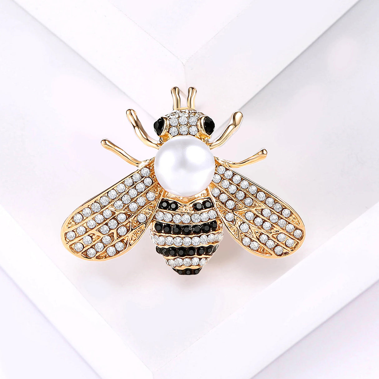 Bee brooch in gold-tone metal with clear faceted stones, black details, and white round bead, flat lay on white background.