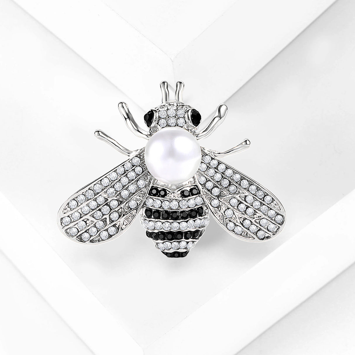 Bee brooch with silver-tone finish, clear faceted stones, black accents, and white round bead, flat lay on white surface.
