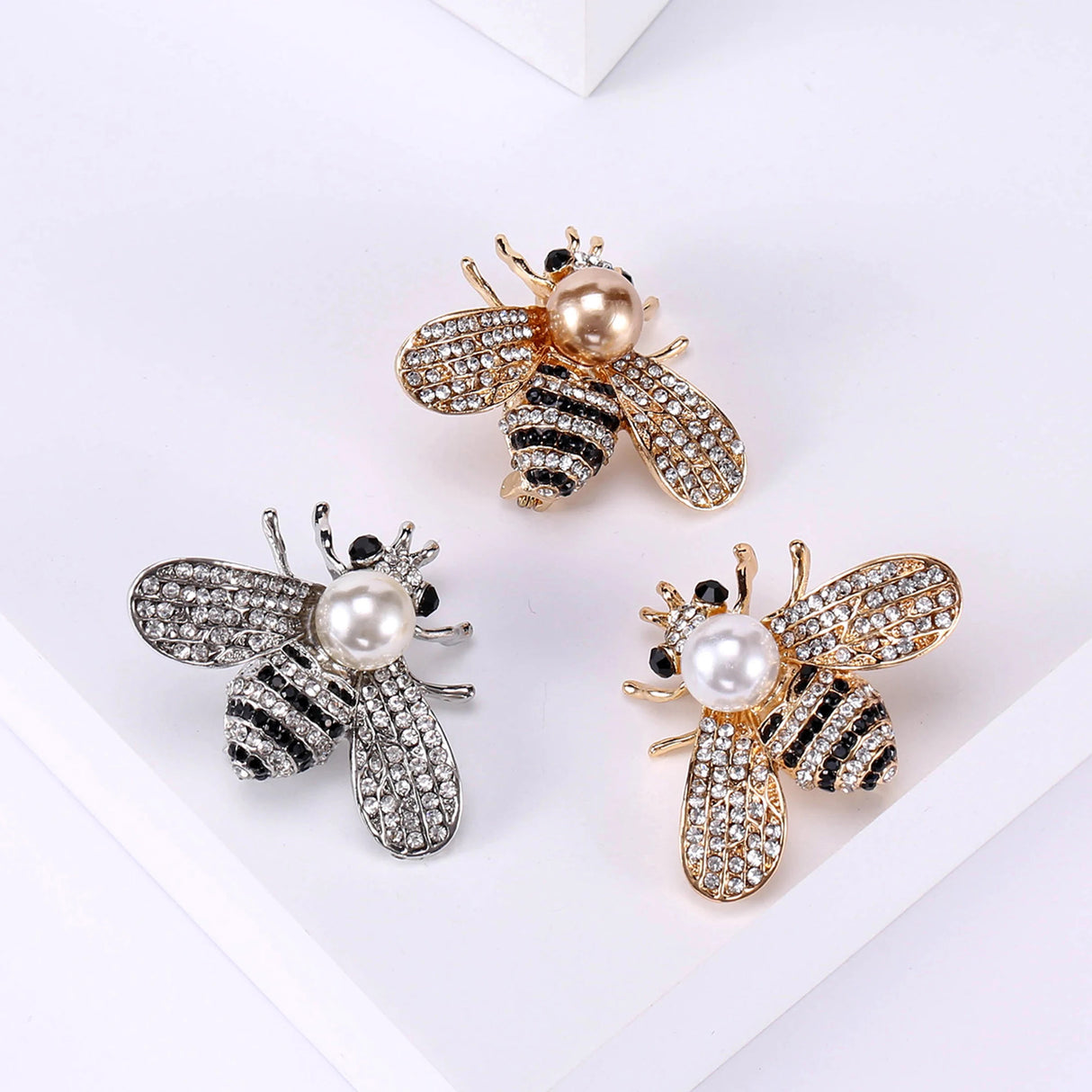 Set of three bee brooches in gold and silver-tone finishes with clear stones, black details, and round beads, flat lay on white.