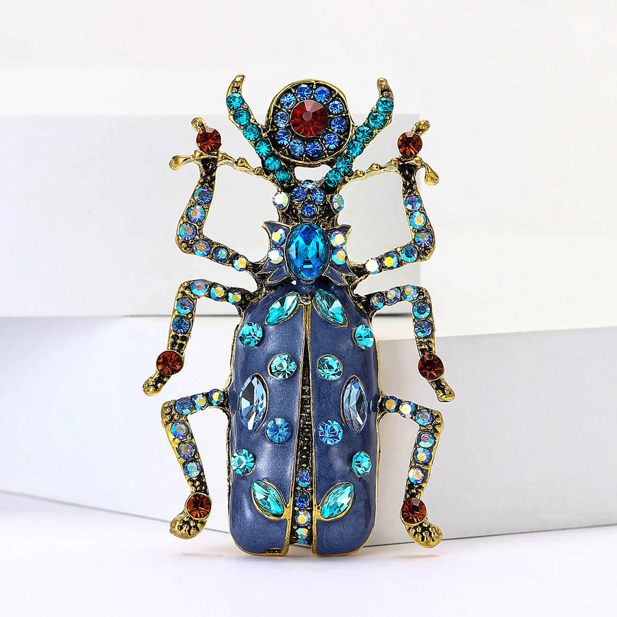 Beetle brooch featuring blue enamel body with gold-tone legs and blue and red stones on wings and antennae, upright front view.