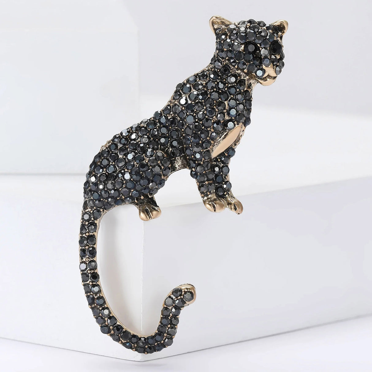 Panther-style brooch with gold-tone base and glossy black rhinestones. Sculptural big cat pose with curled tail, shown upright on white geometric blocks. Ideal as leopard or jungle cat accessory.