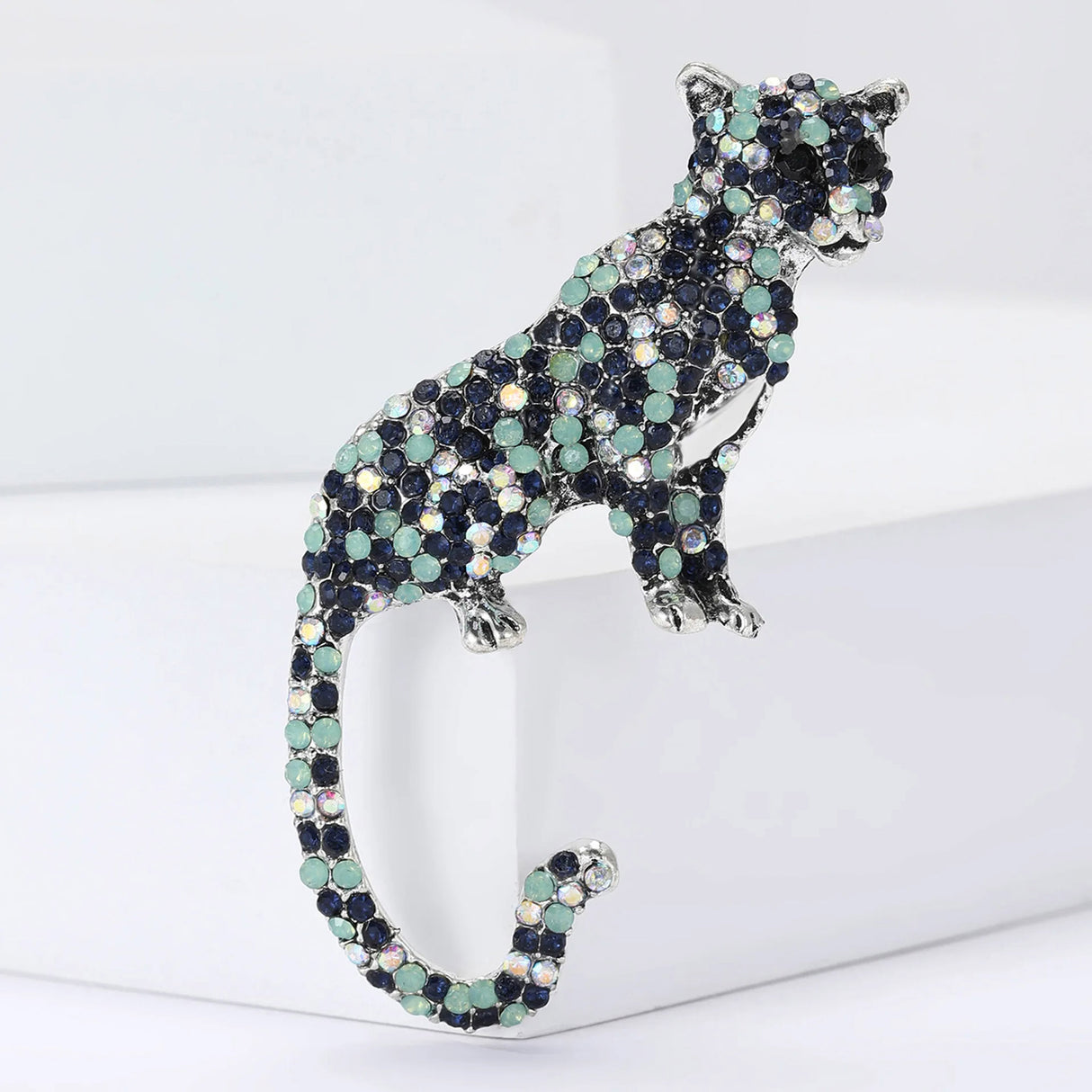Leopard or cheetah-inspired brooch in silver-tone metal, featuring a mix of navy, mint green, and iridescent rhinestones. Upright feline pose with curled tail, styled on clean white block display.