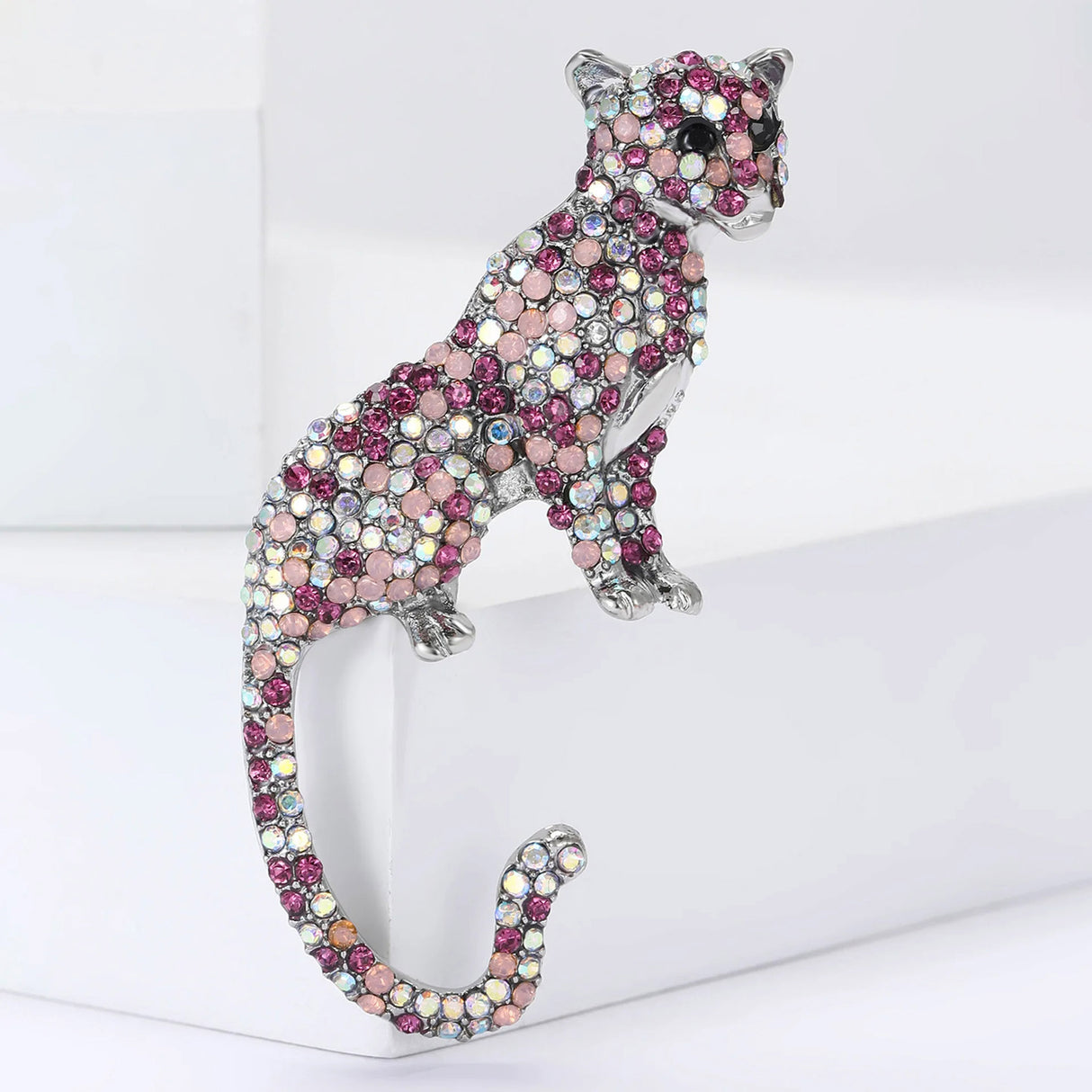 Cheetah-style brooch with silver-tone finish and faceted stones in pink, fuchsia, and shimmering opal. Upright posture with long, curved tail, positioned on a white geometric surface.