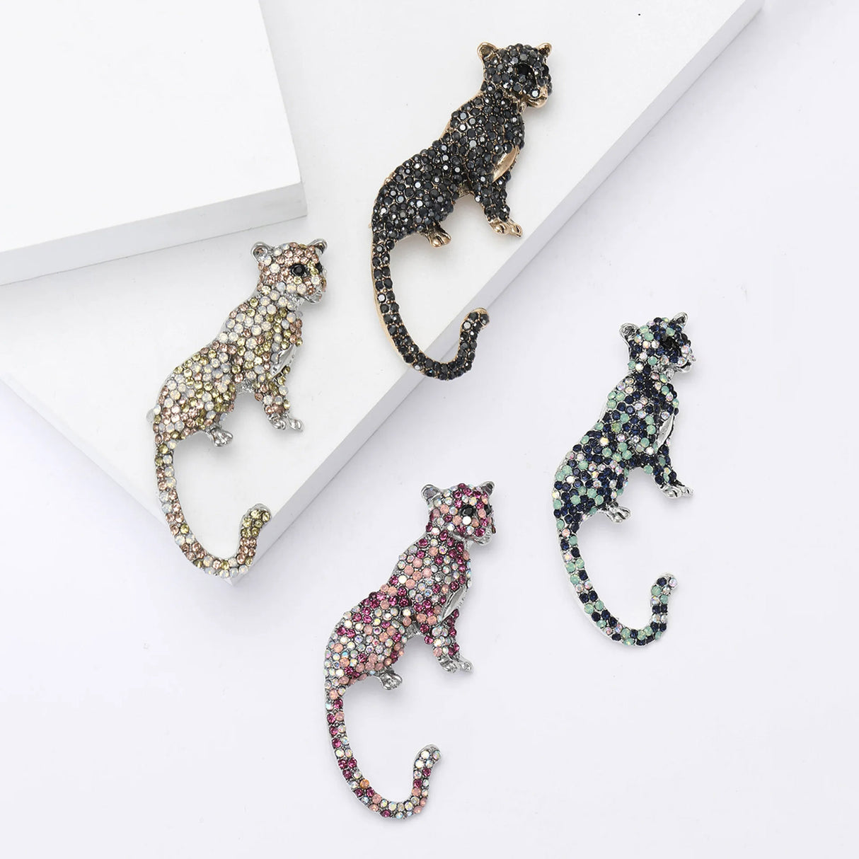 Set of four big cat brooches including panther, cheetah, and leopard styles, in multicolor rhinestones with gold-tone and silver-tone bases. Flat lay on white surface with geometric blocks.