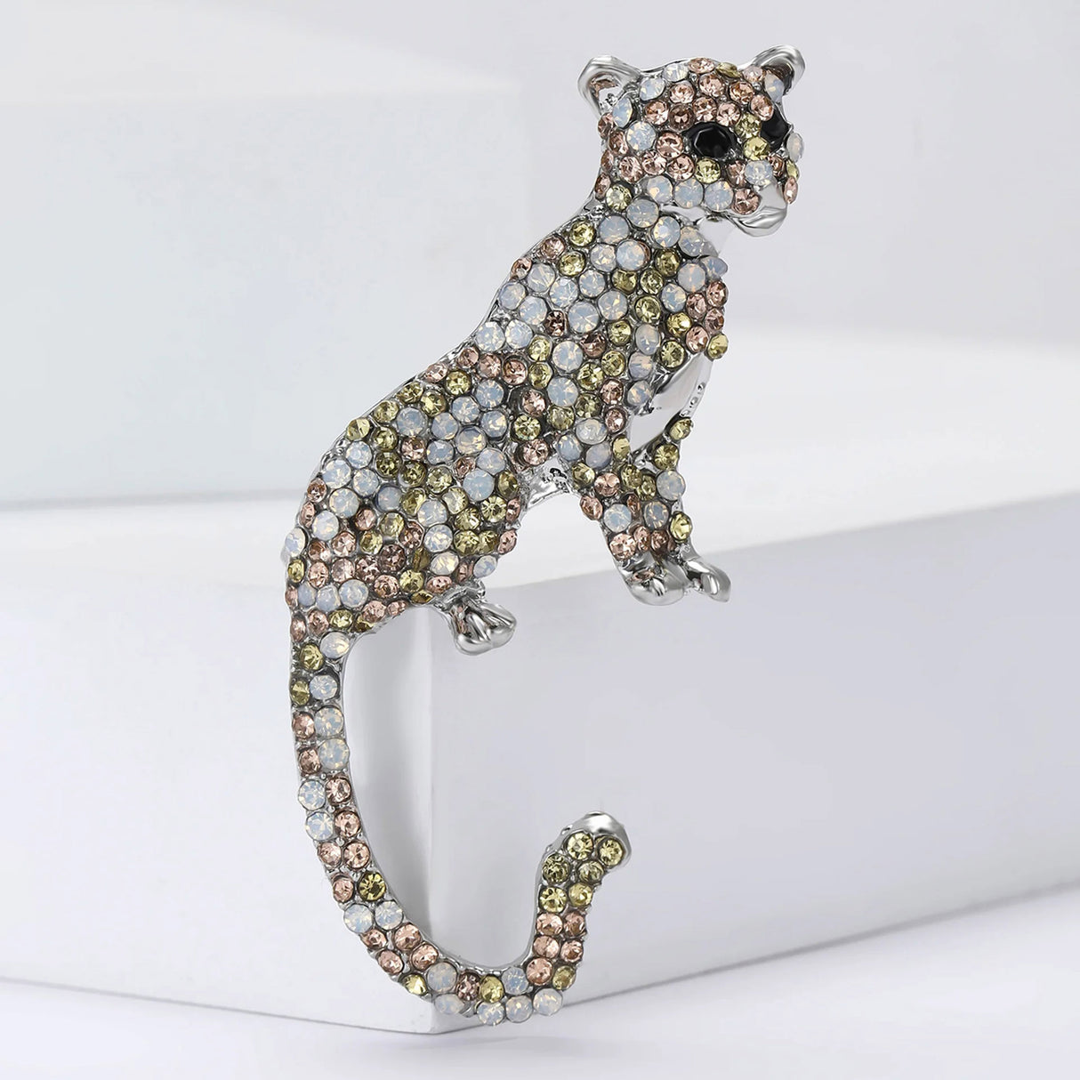 Leopard brooch in silver-tone metal, encrusted with pale champagne, iridescent white, and soft gold rhinestones. Elegant big cat silhouette with curved tail, displayed upright on white display blocks.