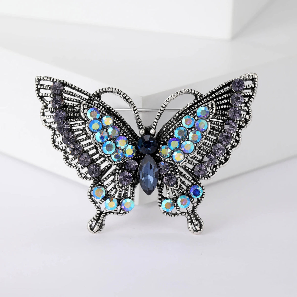 Butterfly brooch with dark silver-tone metal finish, blue faceted center, iridescent black-blue accent stones, front view on white block.