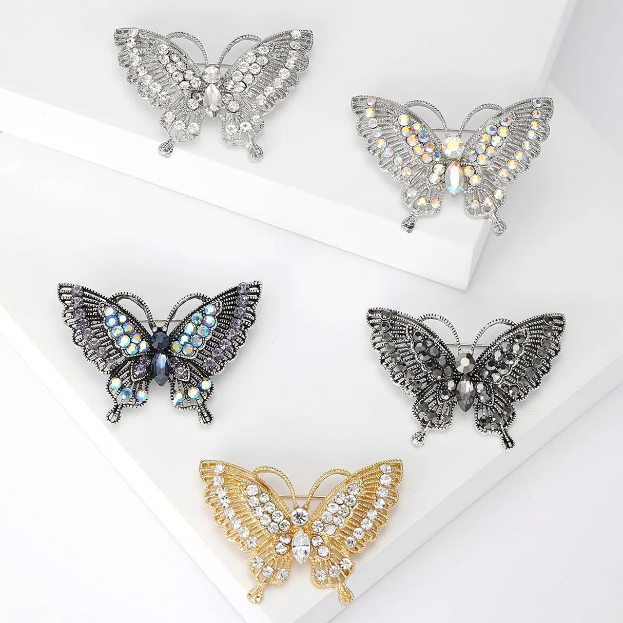 Set of five butterfly brooches in assorted silver, gold, and black tones with clear, iridescent, and dark stones, flat lay on white.