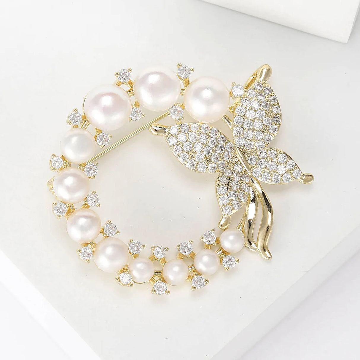 Butterfly brooch with gold-tone metal, clear faceted stones, and white round faux pearls, flat lay on white surface.