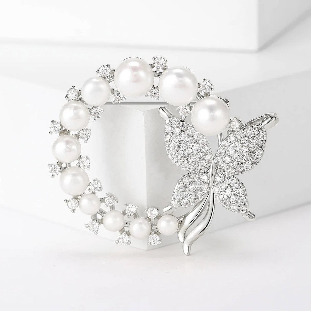Silver-tone butterfly brooch with round white imitation pearls and clustered clear stones in circular layout, standing on white cube.