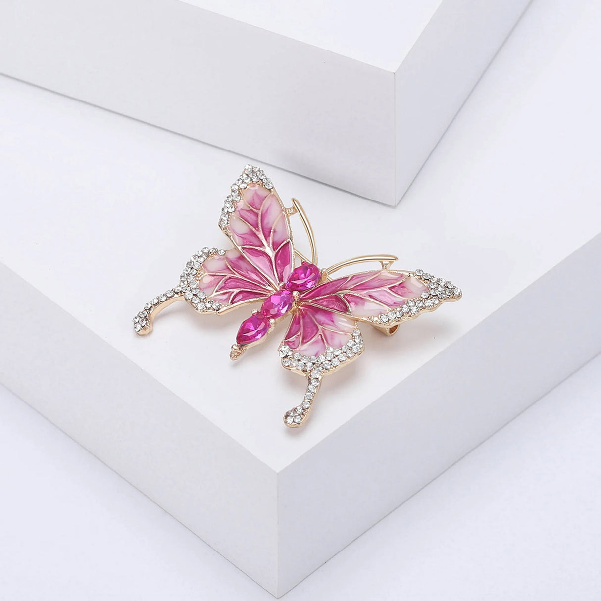 Butterfly brooch with pink enamel wings, gold-tone outline, clear edge stones, and three large vivid pink faceted ovals in the body, angled pose on white platform.