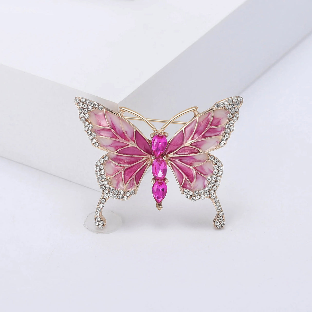Butterfly brooch with pink enamel wings trimmed in gold-tone metal, clear decorative stones, and three pink faceted ovals vertically aligned as body. Upright view on white in studio setting.