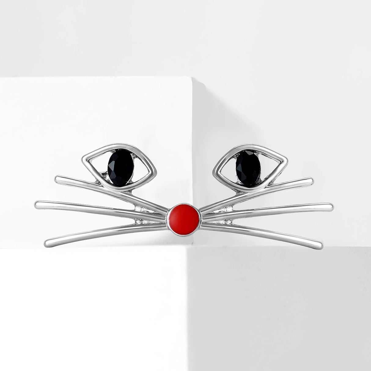 Cat face brooch in silver-tone metal with black oval stones for eyes and red round enamel nose, displayed on white block.