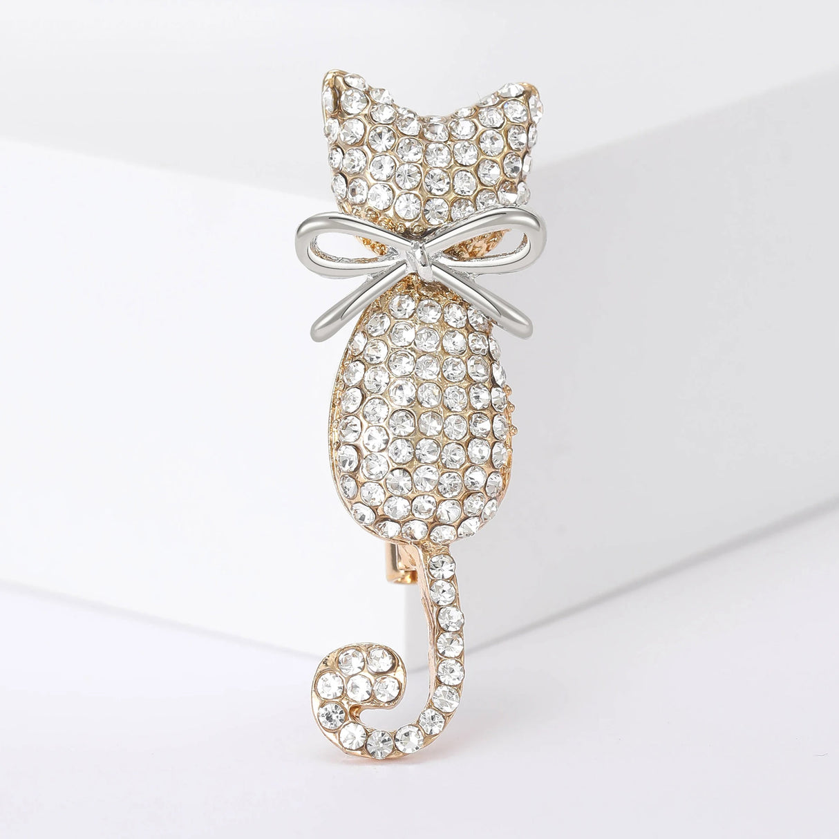 Cat-shaped brooch covered in pave of clear round stones, set in gold-tone metal base, with silver-tone bow. Upright view on white studio background.