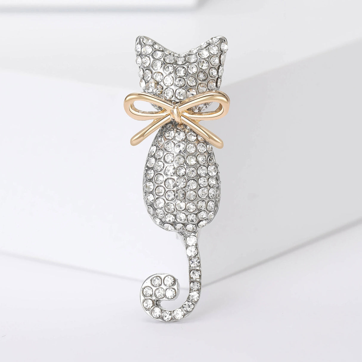 Cat brooch with silver-tone metal base, body covered in small clear round stones, and a gold-tone bow. Standing angled pose on white studio backdrop.