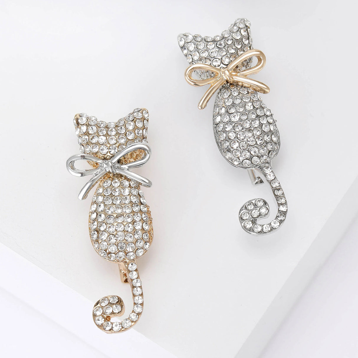 Pair of cat brooches with clear round stone pave, one in gold-tone with silver bow and one in silver-tone with gold bow, flat lay on white.
