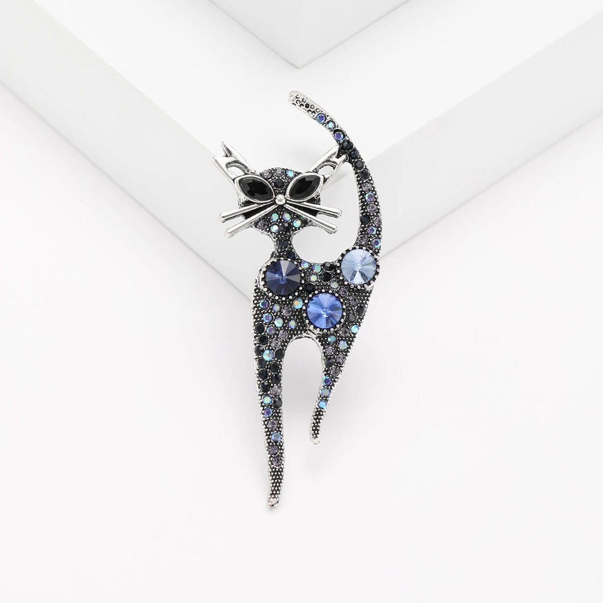 Cat brooch covered in round blue-toned stones, set in vintage style silver-tone metal base, displayed on white platform in studio.