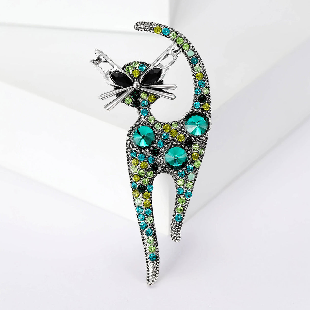 Cat brooch with silver-tone outline, green-toned stones, and large round faceted accents, upright on white platform