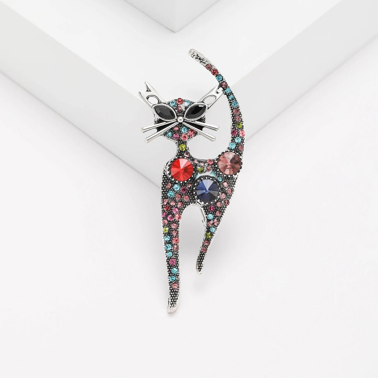 Cat brooch with multicolor faceted stones, silver-tone outline, and black oval eyes, upright on white platform.