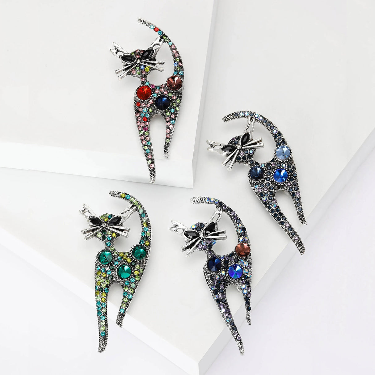Set of four cat brooches with antique-style silver-tone outlines and multicolor faceted stones, arranged on layered white platforms in studio.