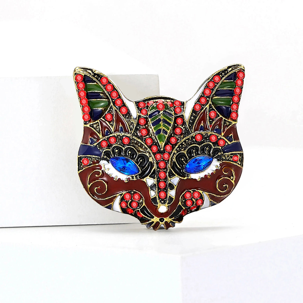 Cat mask brooch with red enamel finish, blue marquise stones for eyes, and multicolor accents including green, blue and black. Upright front view on white background.