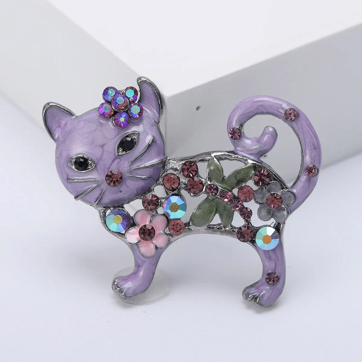 Cat brooch with purple enamel, pink stones, floral details, and green leaf accents, upright pose on white studio backdrop.