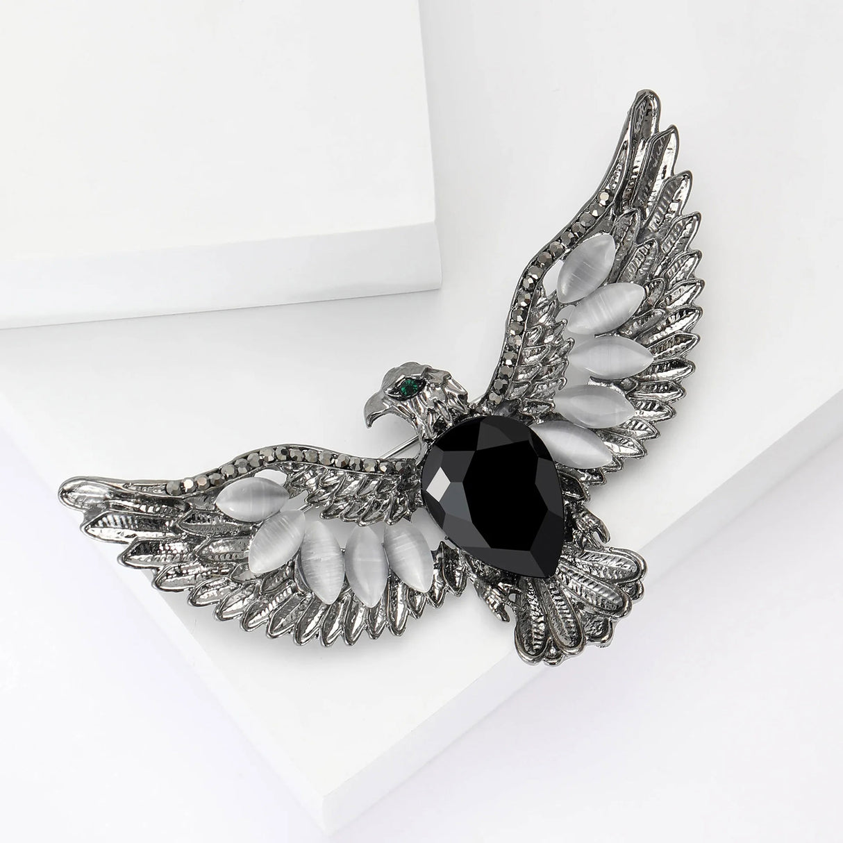 Eagle brooch with silver-tone metal, black faceted center stone, white oval beads, and green eye accent, angled view from top on white surface.
