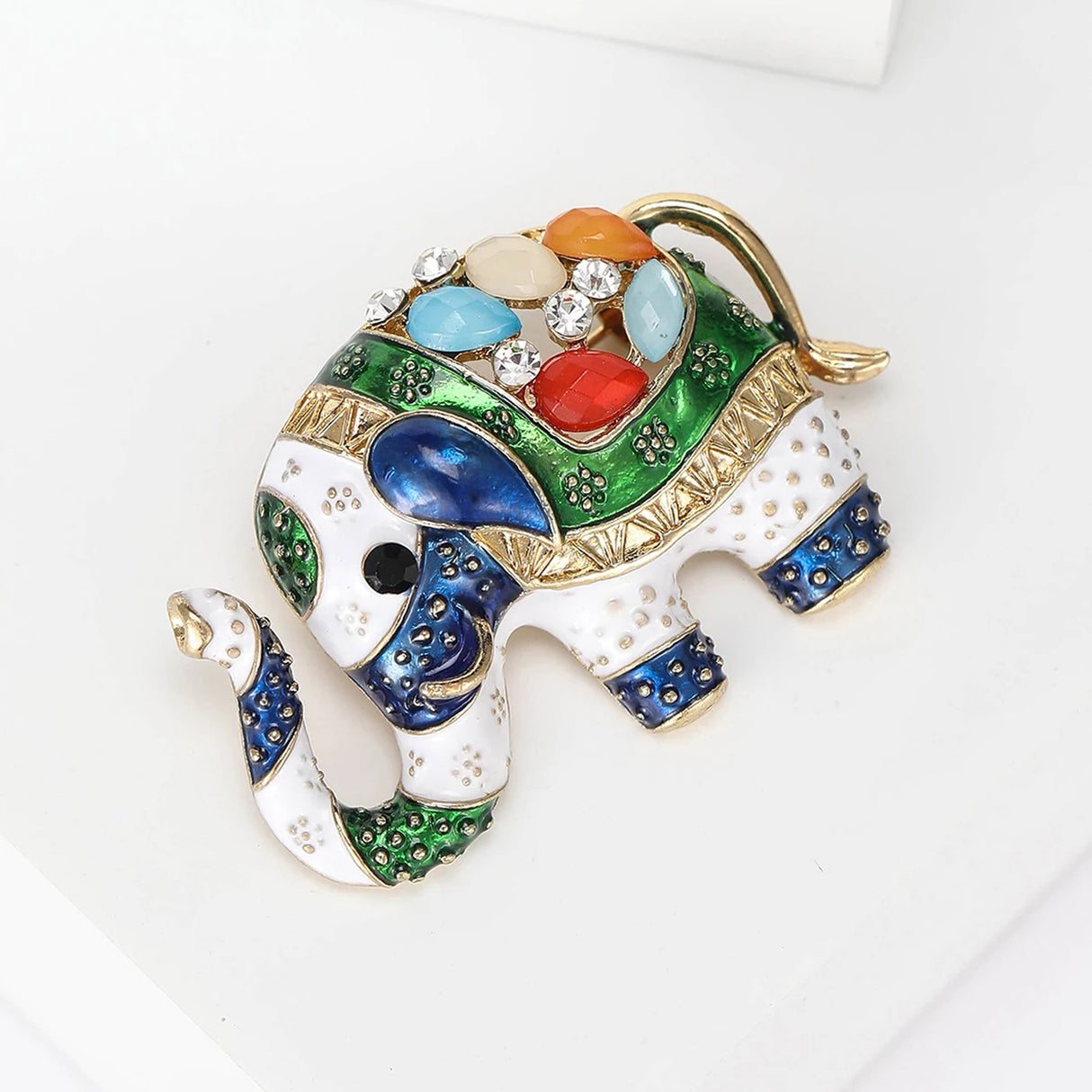 Elephant brooch with green, blue, and white enamel accents, multicolor faceted stones, and gold-tone detailing, angled top-view on white.