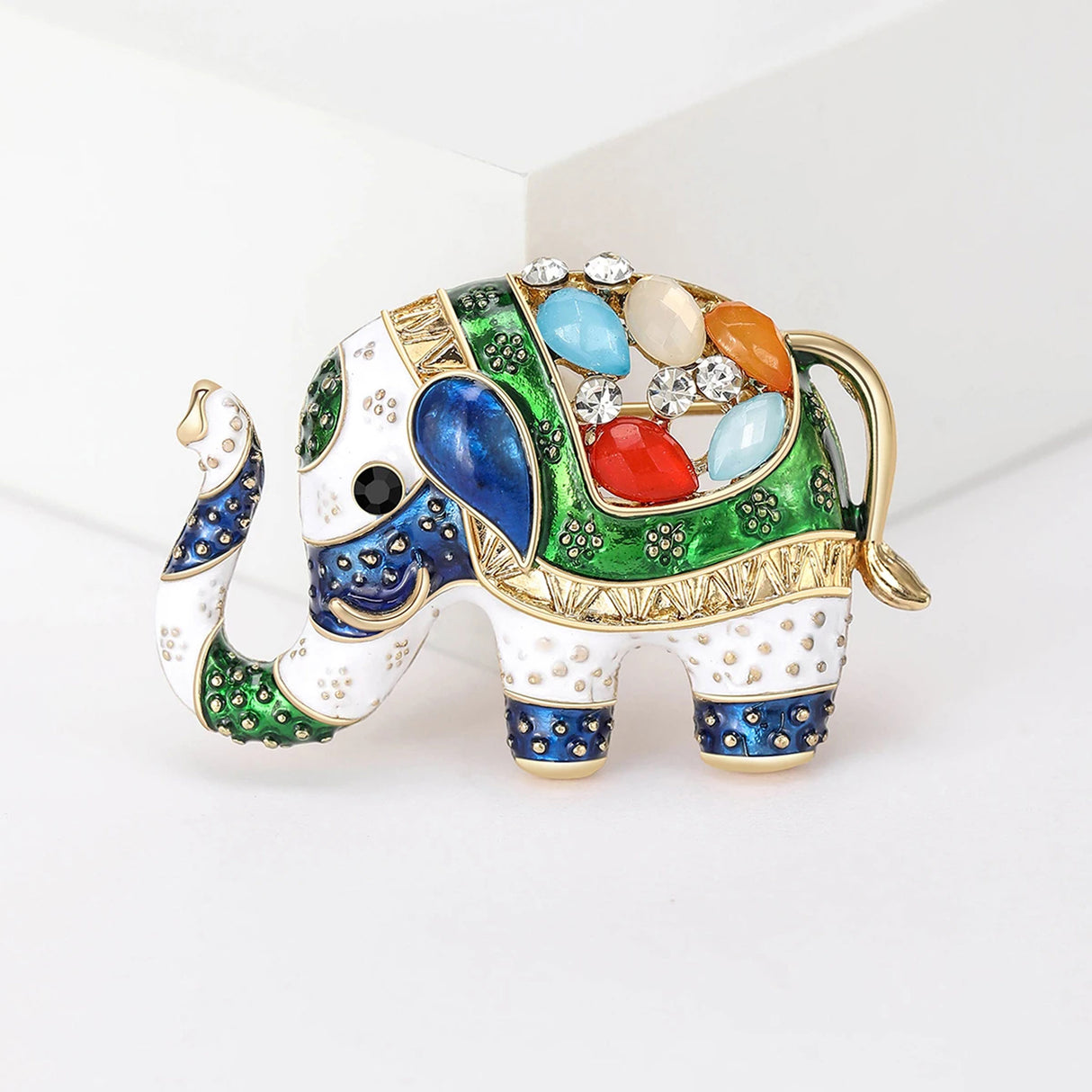 Elephant brooch featuring green and blue enamel, white base with gold-tone trim, and multicolor decorative stones, front studio view.