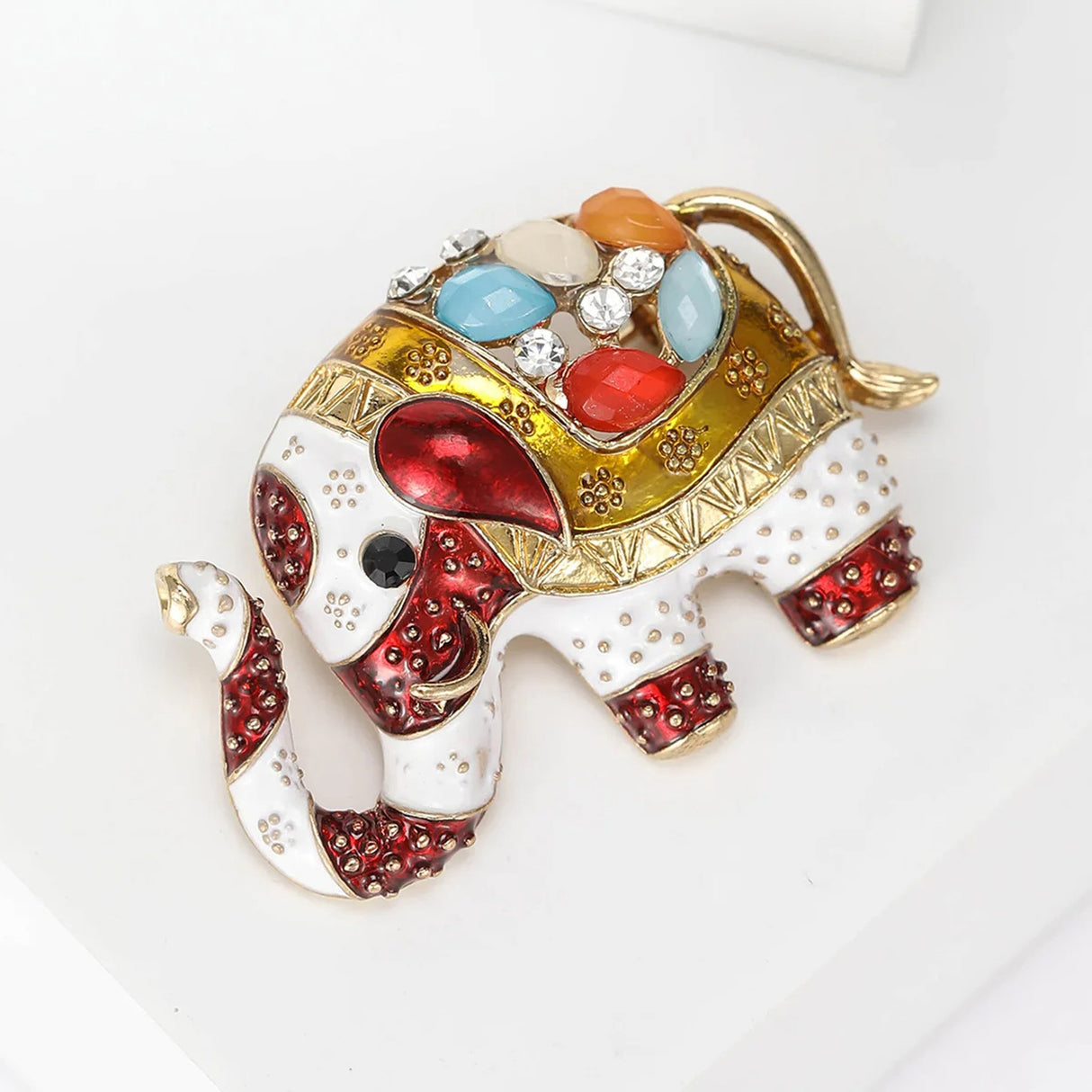 Elephant brooch with red, gold-tone, and white enamel accents, multicolor faceted stones, and textured detailing, angled bird eye's view on white.