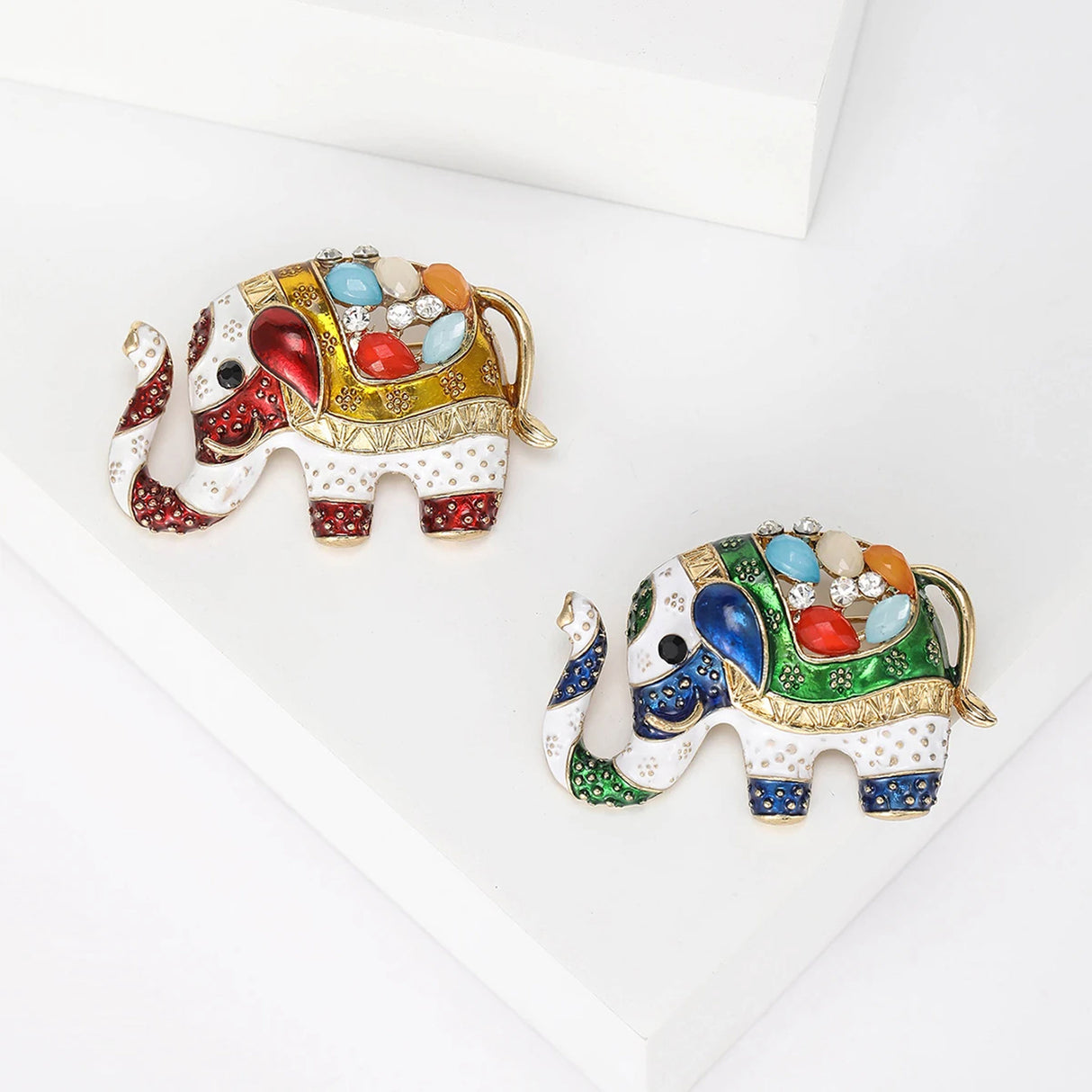 Pair of elephant brooches in red-yellow and blue-green enamel variations with gold-tone details and multicolor faceted stones, flat lay on white background.