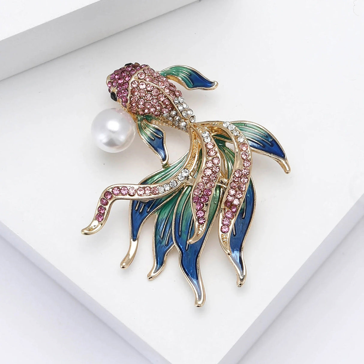 Fish brooch with pink faceted stones, green and blue enamel fins, clear accents, and faux pearl, flat lay on white.