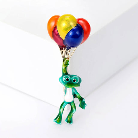 Front view of frog brooch with green enamel body and metallic red, yellow, blue, and orange balloons, staged front view on white studio backdrop.