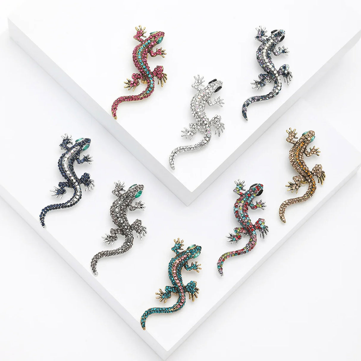 Set of eight lizard brooches in assorted colors including pink, blue, gold, and silver, arranged on white tiered studio display.